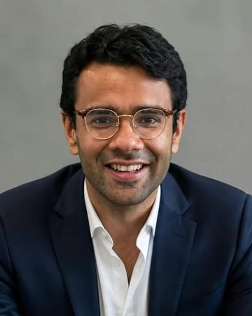 Portrait of Mr. Tanish Broker, smiling, wearing round glasses and a navy blazer over an open white shirt